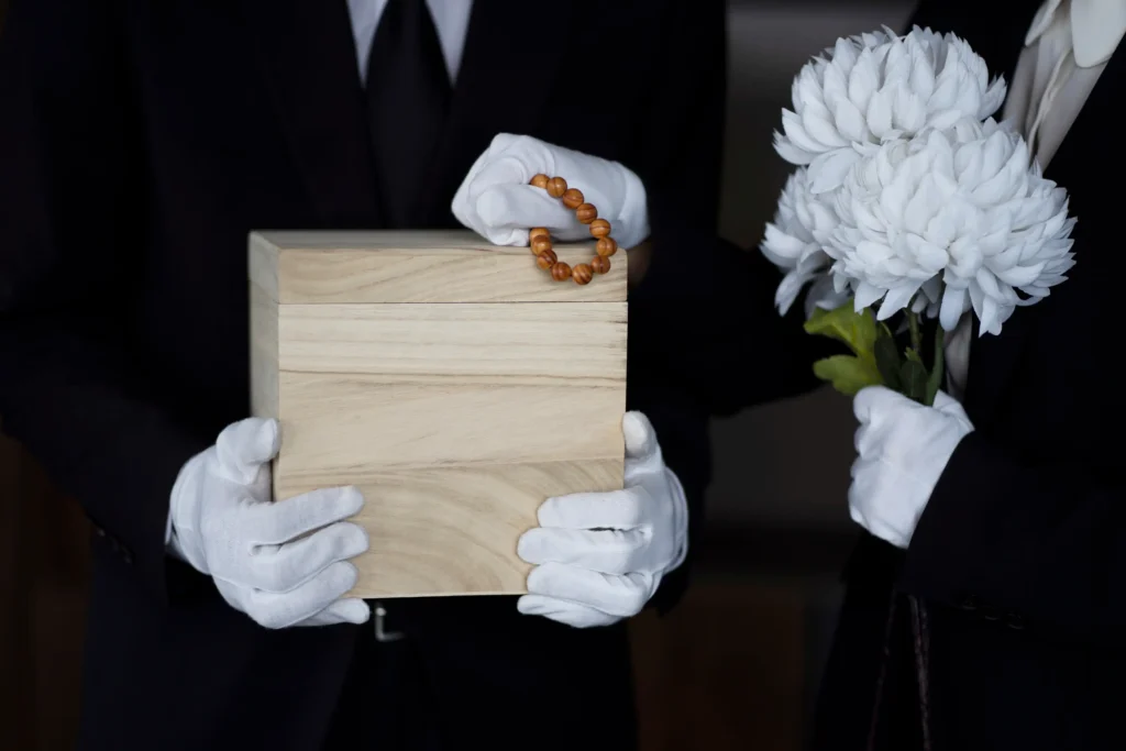 Officials in black suits and white gloves holding a box-shaped, unfinished wooden urn and large, white flowers