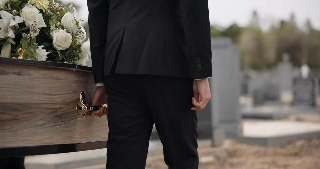 A man in a black suit helping to carry a casket covered in white flowers into a cemetery
