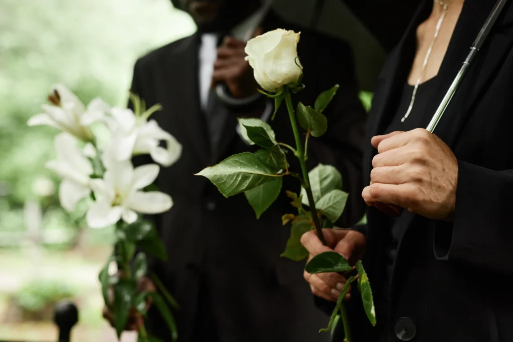 Two mourners holding umbrellas and white flowers at an outdoor funeral service