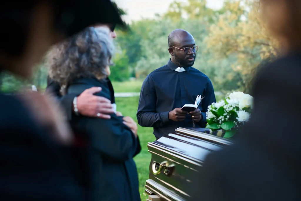 A priest reading from a book at the head of a casket during an outdoor funeral service