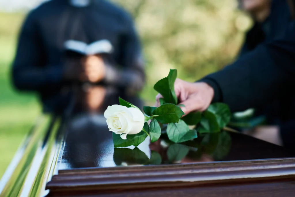 A mourner placing a white rose on a casket while a priest reads from a notebook